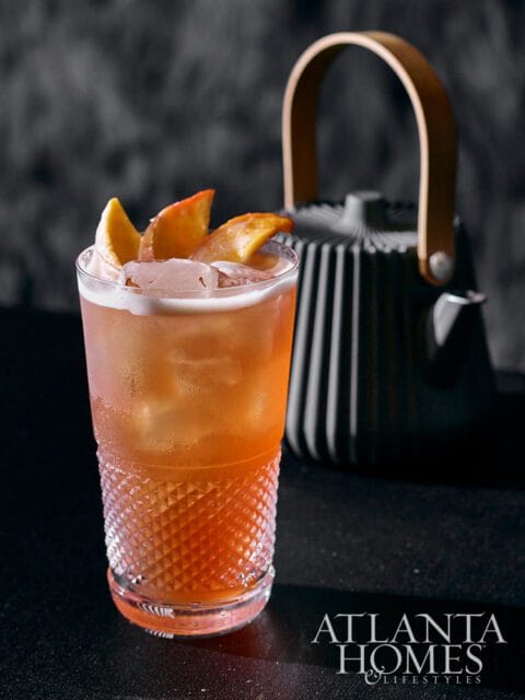 Tall glass with orange cocktail, ice, and peach slices. Black teapot in background.