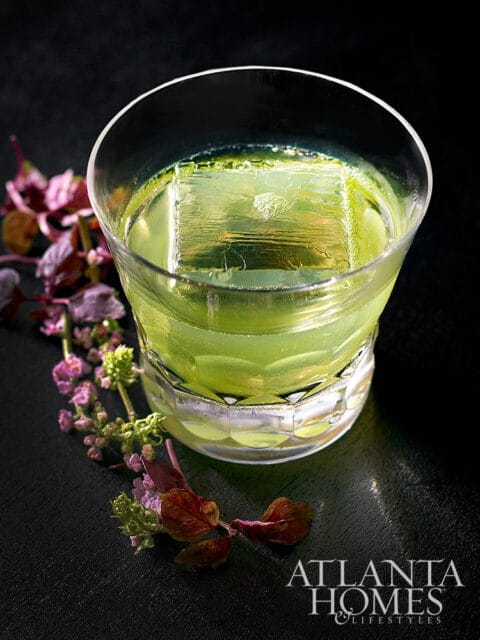 Green cocktail with a large ice cube and floral garnish, Atlanta Homes & Lifestyles logo