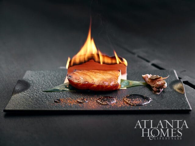 Seared fish on a black plate with a wooden block on fire, garnished with a small piece of food and