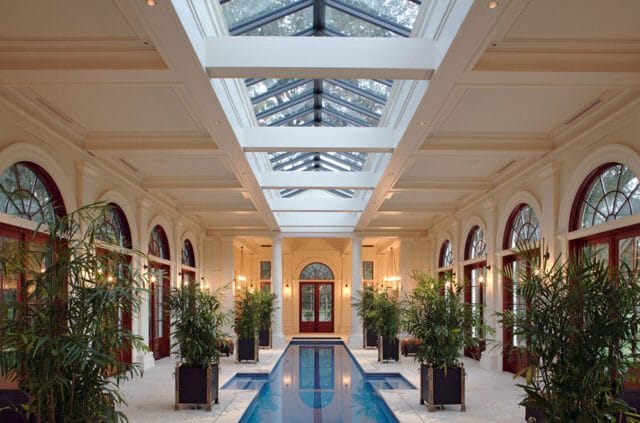 Indoor lap pool with glass ceiling, arched windows, and potted plants.