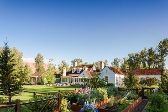 White house with red tile roof and stone chimneys, surrounded by trees and a garden.