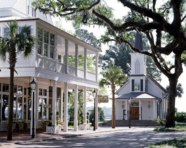 White building with a steeple and wrap-around porch, surrounded by trees and palm trees.