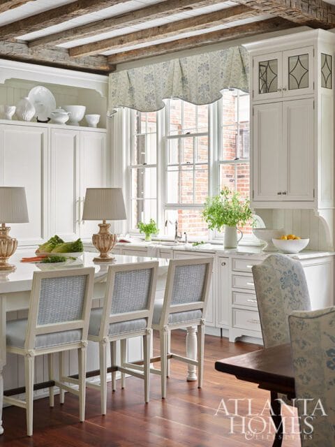 White kitchen with blue floral valance, island with bar stools, and exposed wood beams.