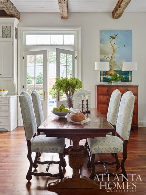 Dining room with wooden table, upholstered chairs, French doors, and artwork.