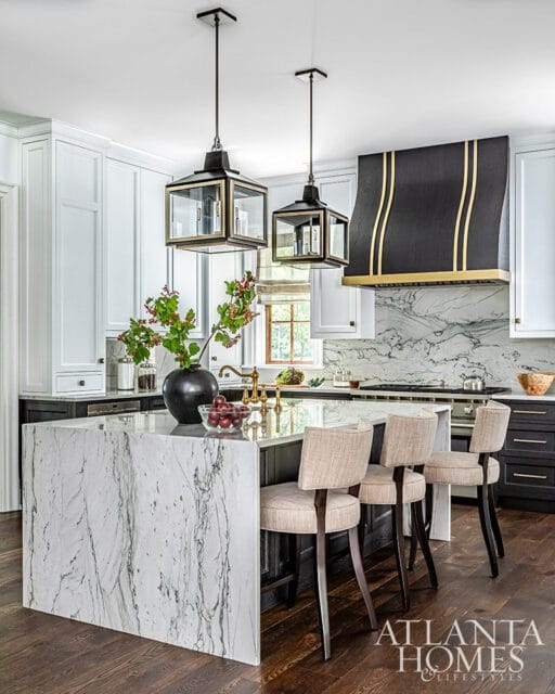 Kitchen island with marble waterfall edge, pendant lights, and black range hood with gold accents.
