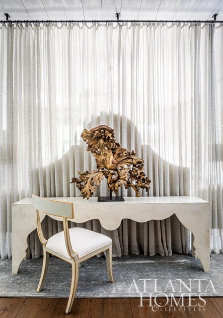 Cream console table with ornate gold sculpture and upholstered chair in front of sheer curtains.