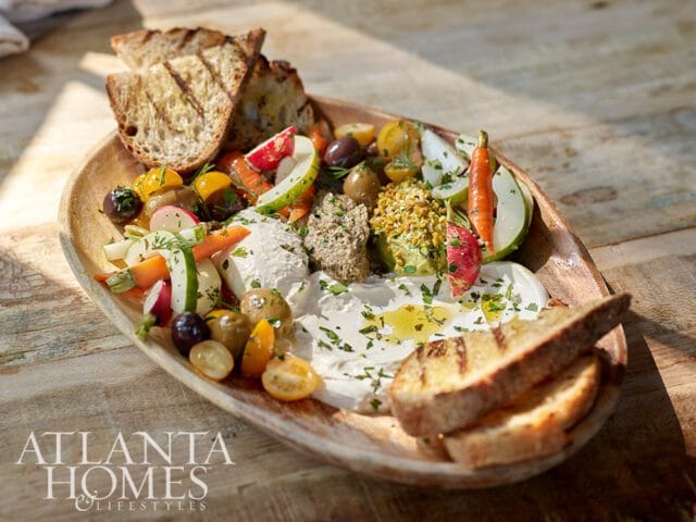 Wooden platter with dips, grilled bread, olives, cherry tomatoes, and sliced vegetables