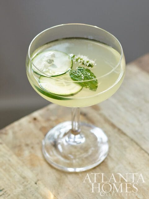 Cocktail with cucumber slices, mint, and small white flowers in a coupe glass.