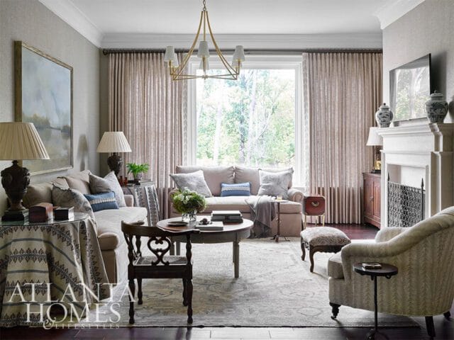 Living room with neutral sofas, fireplace, ornate chandelier, and large window.