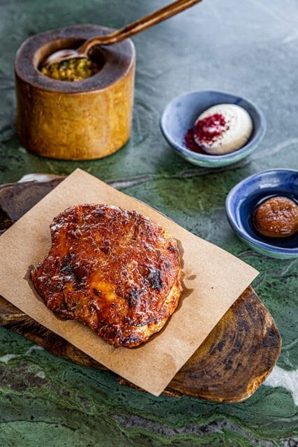 Roasted chicken thigh with crispy skin, served on brown paper with sides of dip and sauce.