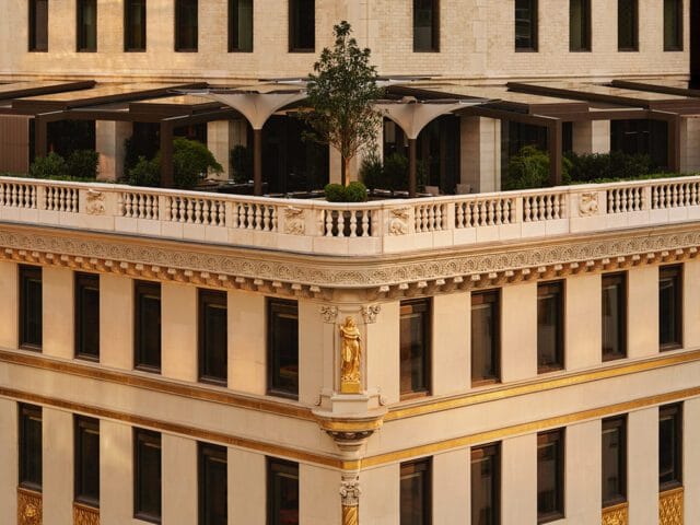 Cream building with ornate gold trim and a statue on the corner. Balcony with railing and trees