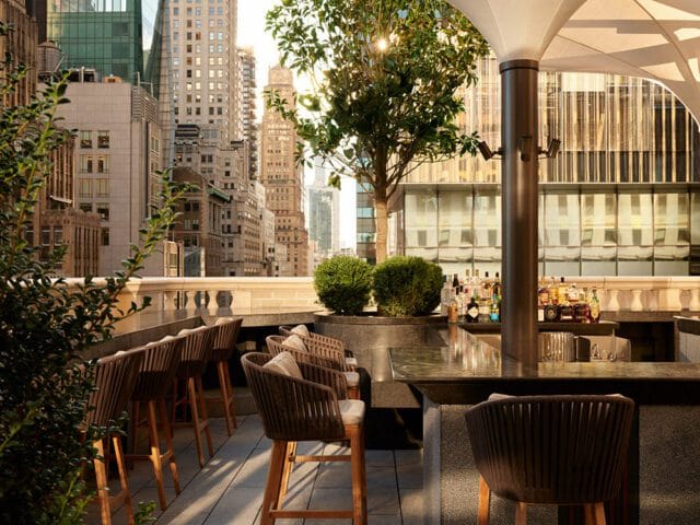 Rooftop bar with city skyline view, seating, and bar stocked with bottles.