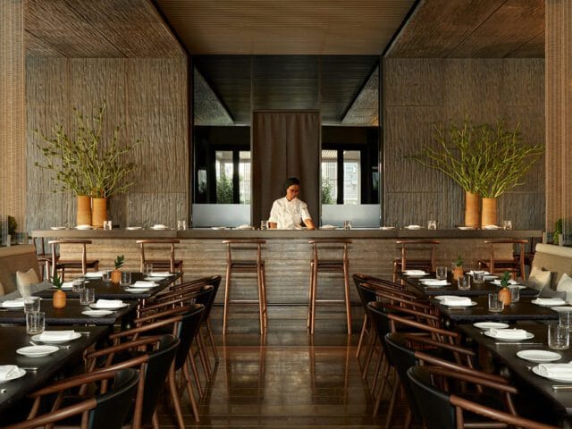 Restaurant dining room with wood tables, chairs, and a bar. A chef stands behind the bar.