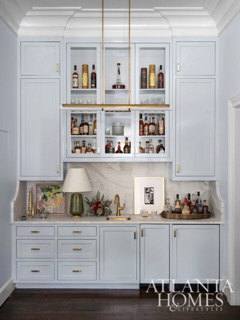 Light blue bar with glass-front cabinets filled with bottles, marble backsplash, and brass hardware.