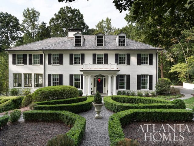 White brick house with black shutters and a gray roof, surrounded by manicured hedges.
