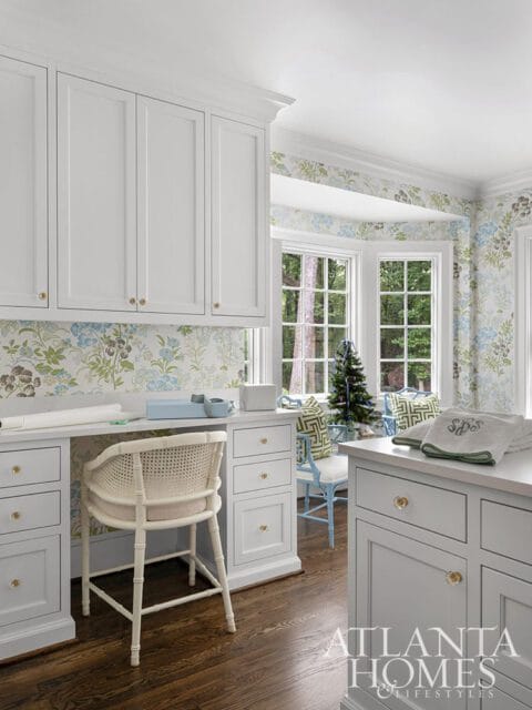 White built-in desk with cane chair, floral wallpaper, and bay windows.