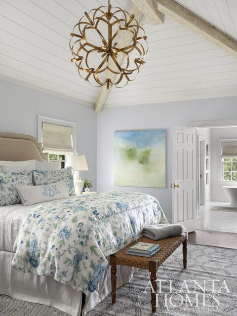 Bedroom with vaulted ceiling, floral bedding, and brass chandelier. Open doorway reveals bathroom.