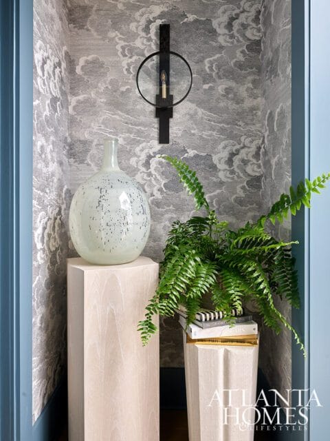 Light wood pedestal with speckled vase, fern on books, and sconce on cloud wallpaper.
