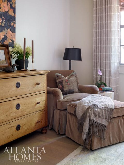Brown armchair and ottoman with a fringed throw, next to a wooden dresser with decorative hardware.