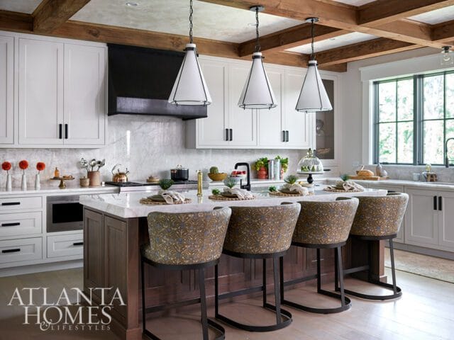 White kitchen with marble island, wood beams, and upholstered bar stools.