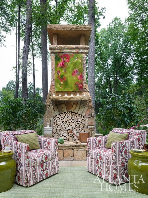 Two armchairs upholstered in floral fabric flank a stone fireplace with stacked firewood and