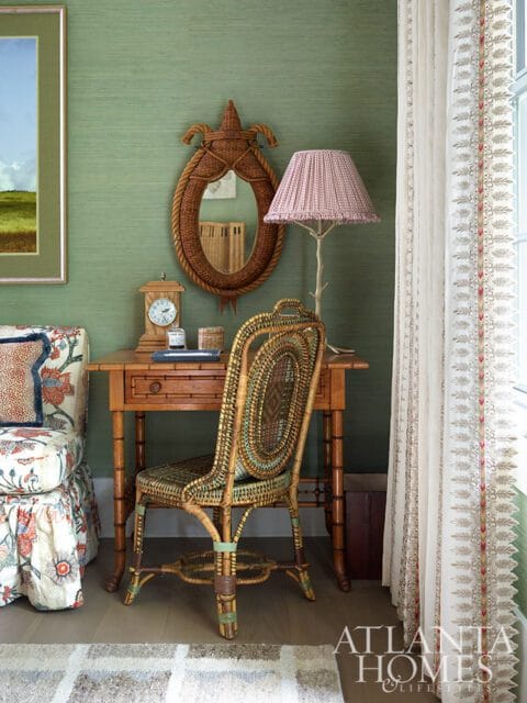 Green grasscloth wallpaper with a wooden desk, wicker chair, and patterned armchair.