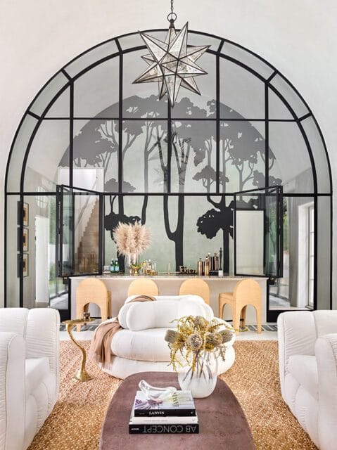 Living room with a large arched window featuring a black tree silhouette mural, a bar area with rattan stools, and white tufted sofas.
