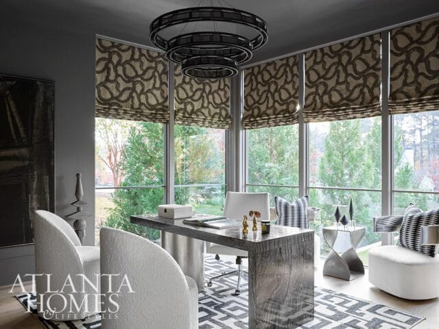 Modern office with a large wooden desk, white boucle chairs, geometric rug, and floor-to-ceiling windows overlooking a garden.