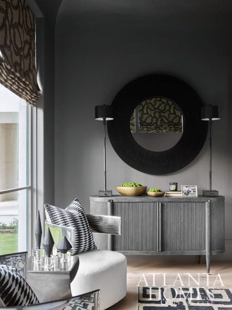 Gray room with fluted console table, large round textured mirror, two black lamps, and a white ottoman with black and white patterned pillows.