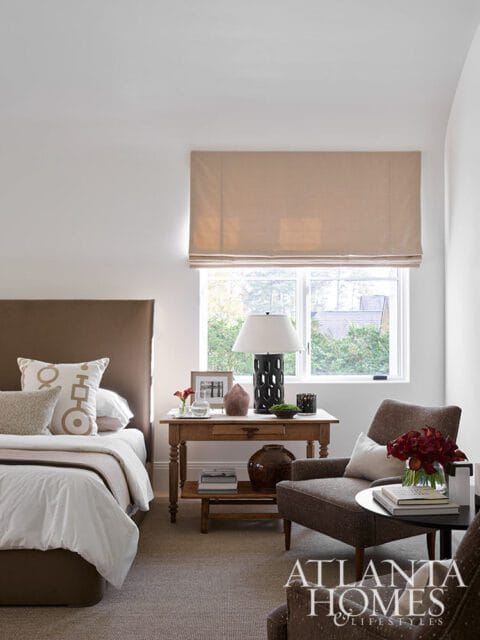 Bedroom with a brown upholstered headboard, white bedding, and decorative pillows. A wooden side table with a lamp and vase sits next to a window with a tan roman shade. A brown armchair with a small round table is in the foreground.