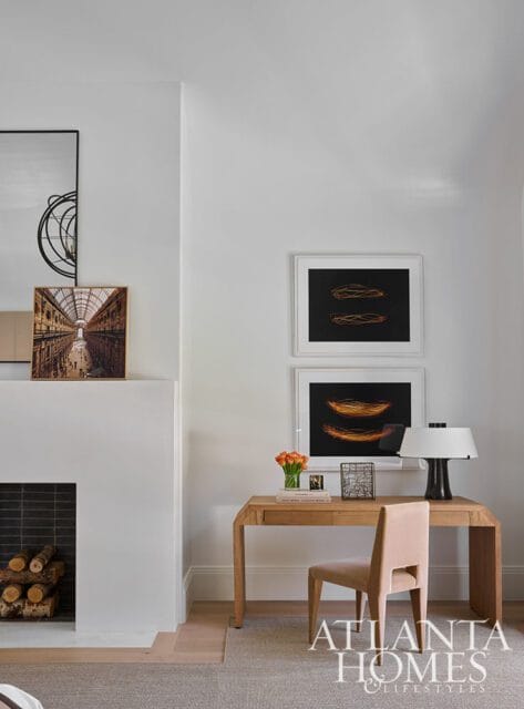 Modern home office with a light wood desk, blush velvet chair, and abstract art above the desk. A white fireplace with stacked logs is to the left.