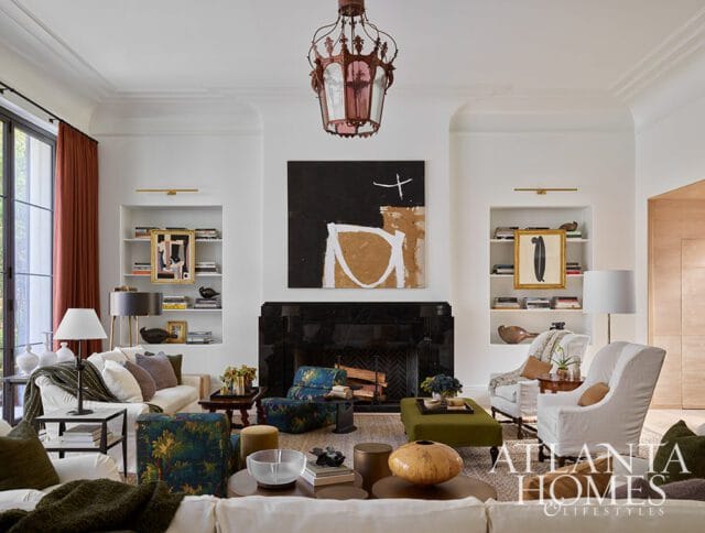 Living room with white sofas, patterned armchairs, abstract art above a black fireplace, and a pink ornate chandelier.