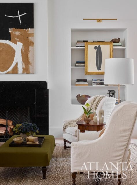 Living room with a black marble fireplace, abstract art, built-in bookshelves, and two white slipcovered armchairs flanking a small wooden side table.