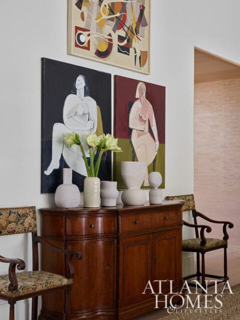Antique wooden sideboard topped with white vases and artwork depicting figures and abstract shapes. Two chairs flank the sideboard.