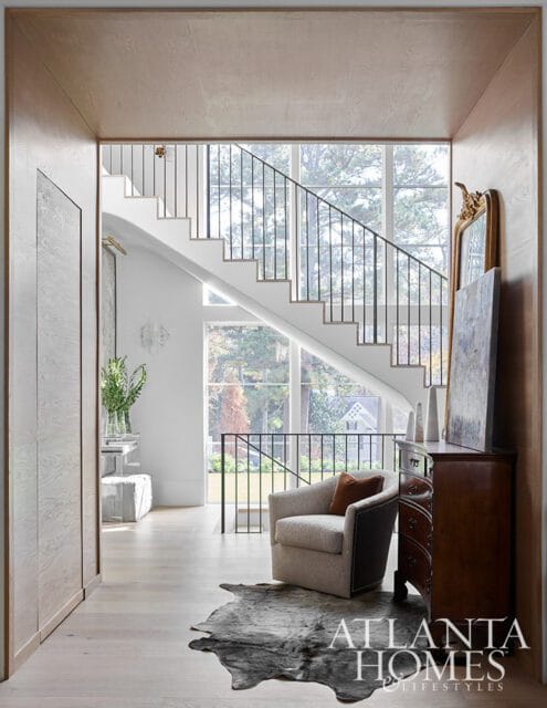 Modern entryway with light wood paneling, a white staircase with black railings, a plush armchair on a cowhide rug, and an antique dresser.