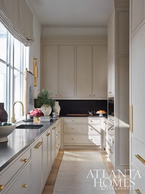 Neutral kitchen with light wood floors, light gray cabinetry, brass hardware, and a dark countertop. A large window with a Roman shade is on the left.