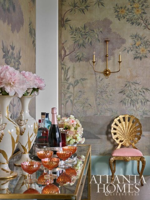 Bar cart with champagne and glassware next to a gold shell-back chair with pink cushion, against a wall with botanical wallpaper and a brass sconce.
