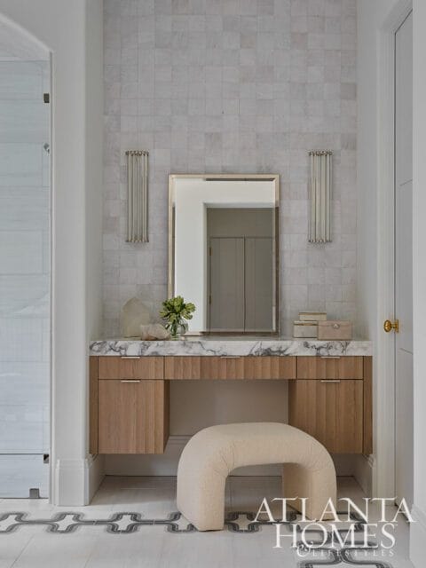 Bathroom vanity with light wood cabinets, marble countertop, and a cream boucle stool. The wall is tiled with small grey squares and features two vertical sconces.