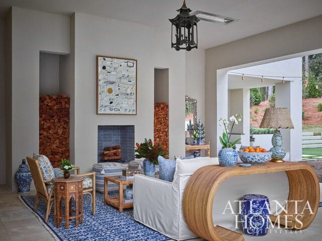 Living room with a fireplace, stacked wood, rattan furniture, blue and white patterned rug, and a curved wooden console table with decorative items.