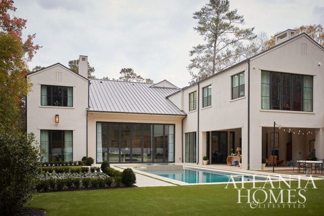 Modern stucco home with a gray metal roof, featuring large black-framed windows, a swimming pool, manicured hedges, and a patio with outdoor seating.