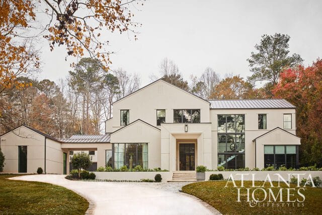 Modern beige stucco house with dark-framed windows and a metal roof, set against a backdrop of autumn trees.