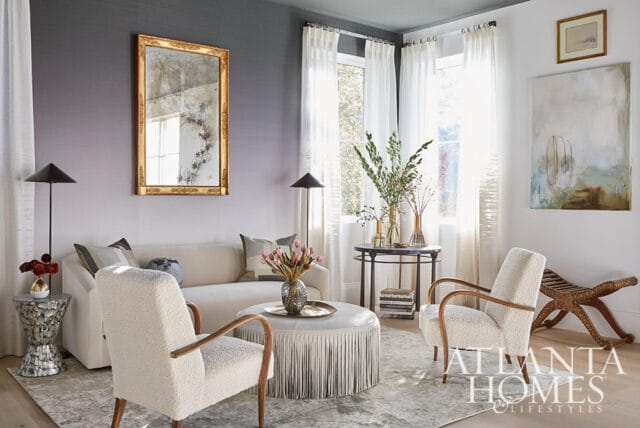 Living room with a cream sofa, two textured armchairs, a fringed ottoman, and a gold-framed mirror on a gray ombre wall.