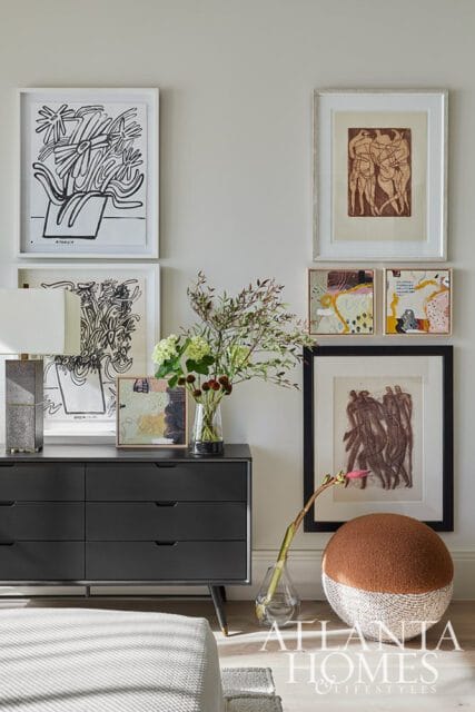 Dark gray dresser with six drawers topped with framed artwork, a lamp, and a vase of flowers. A round ottoman with brown and cream woven texture sits on the floor next to a framed drawing.