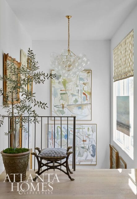 Hallway with abstract art, a glass bubble chandelier, and a decorative stool. A large potted plant sits beside a metal railing.