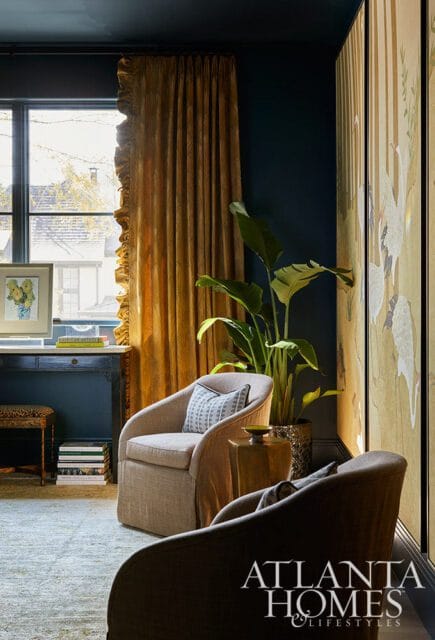 Dark blue walls with gold ruffled curtains framing a window, two upholstered chairs, a large potted plant, and a decorative screen with crane motifs.