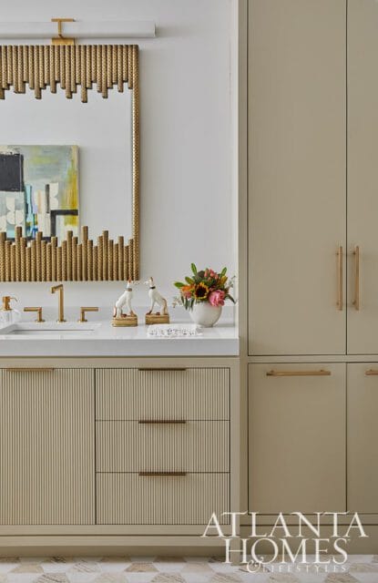 Bathroom vanity with fluted cream cabinetry, white countertop, gold faucet, and a decorative mirror with gold accents. Tall cream cabinets are to the right.