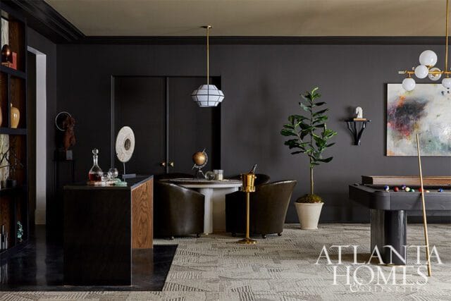 Dark gray room with a pool table, two leather armchairs, a round table with a globe, and a bar area with decanters. A large abstract painting hangs on the wall.