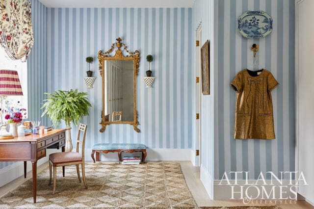 Hallway with light blue and white striped wallpaper, a wooden desk with a fern, a gold ornate mirror, and a gold sequined dress hanging on the wall.