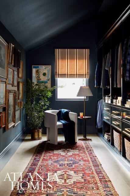 Dark blue walk-in closet with a white armchair, a striped roman shade, and a patterned runner rug on light wood floors.