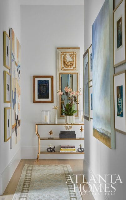 Hallway with abstract art on the left, a console table with decorative objects and books, and a large abstract painting on the right wall. The Atlanta Homes & Lifestyles logo is visible in the lower right corner.